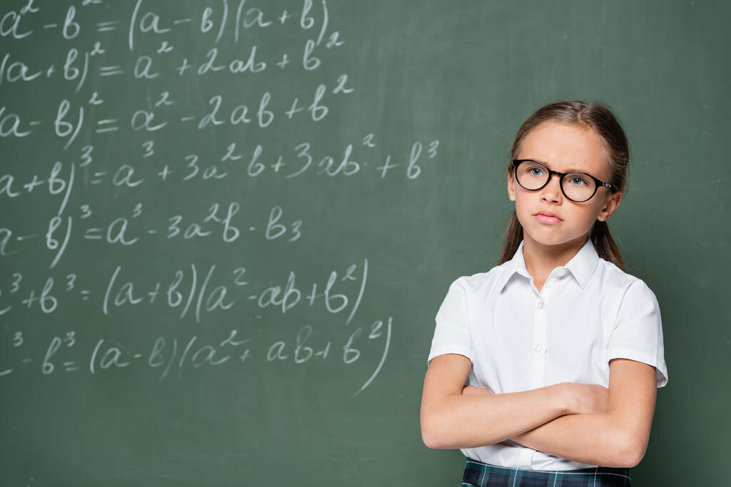 offended schoolgirl standing with crossed arms near chalkboard with equations - Foto, Imagen
