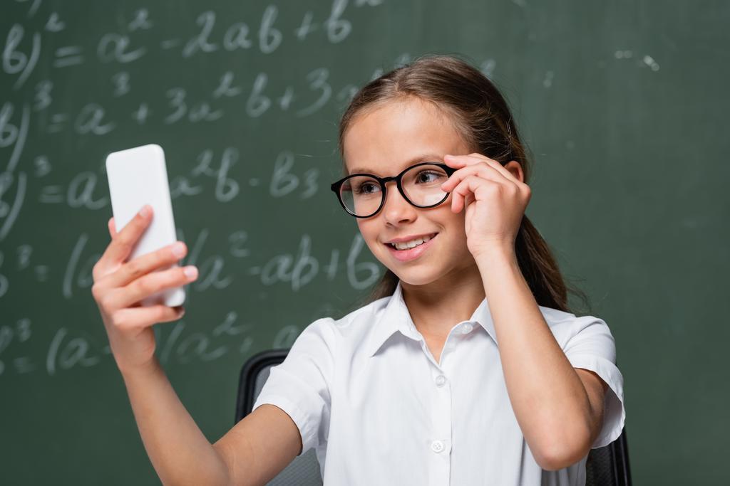 smiling schoolkid touching eyeglasses during video call near chalkboard  - Foto, Imagen
