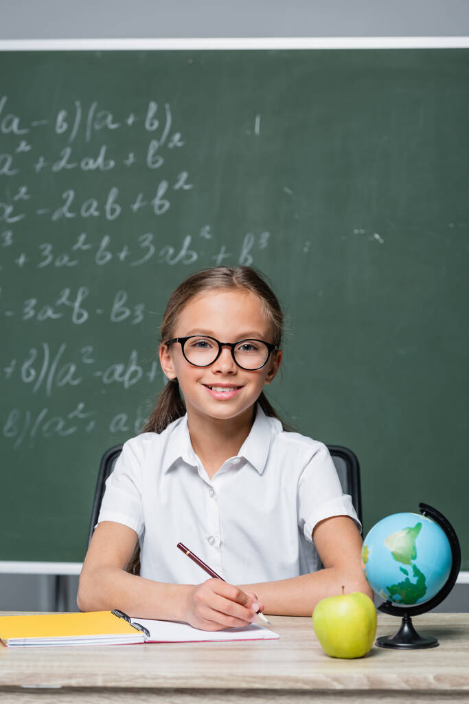 pozitivní školačka s perem s úsměvem na kameru poblíž zeměkoule a notebook na stole - Fotografie, Obrázek