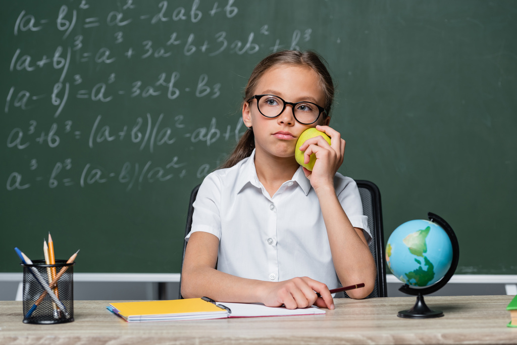 elégedetlen iskolás lány gazdaság alma közelében földgömb és notebook az asztalon - Fotó, kép
