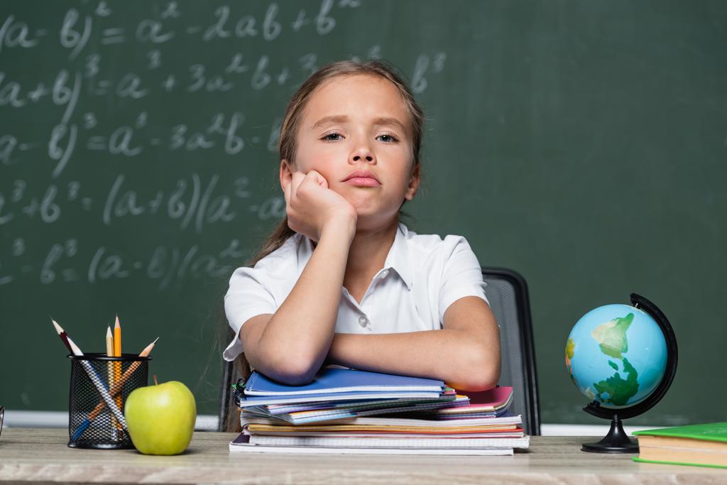 расстроенная школьница смотрит в камеру рядом с блокнотами, глобусом и доской на размытом фоне - Фото, изображение