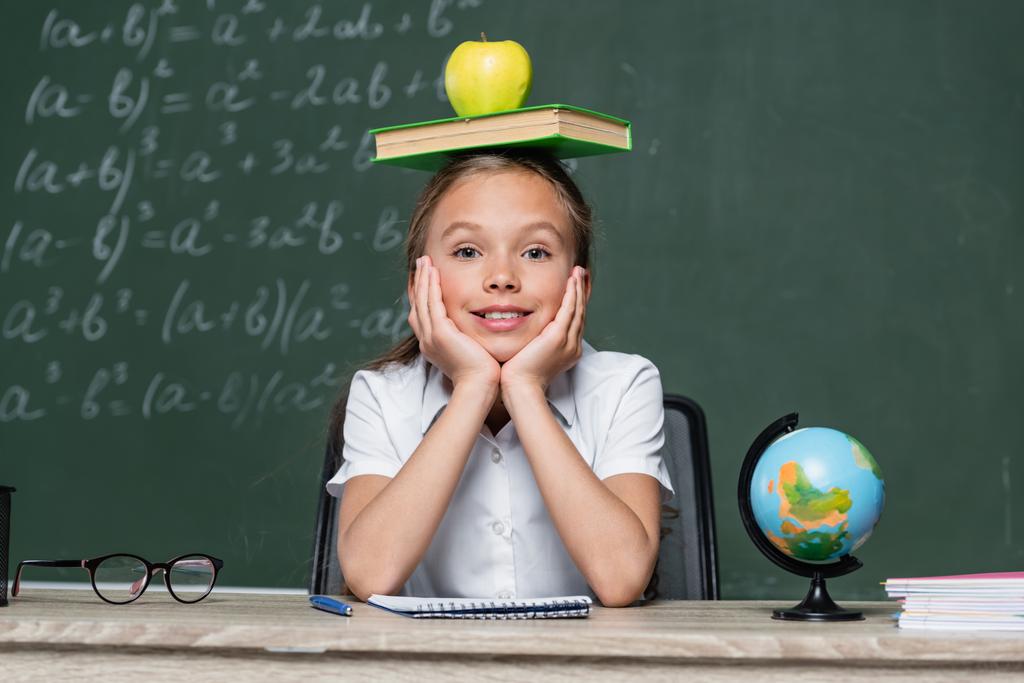 speels schoolmeisje zitten aan het bureau met boek en appel op het hoofd - Foto, afbeelding