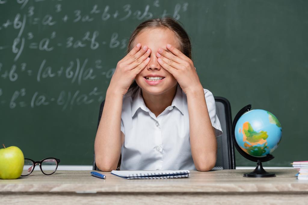 cheerful schoolgirl covering eyes with hands near globe and notebook on desk - Photo, image