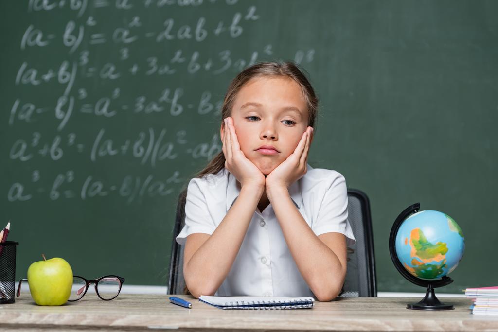 расстроенный школьник сидит за столом рядом глобус, ноутбук и яблоко - Фото, изображение
