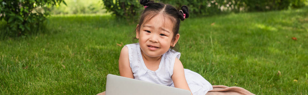 zavaros ázsiai gyerek nézi kamera közelében laptop a parkban, banner - Fotó, kép