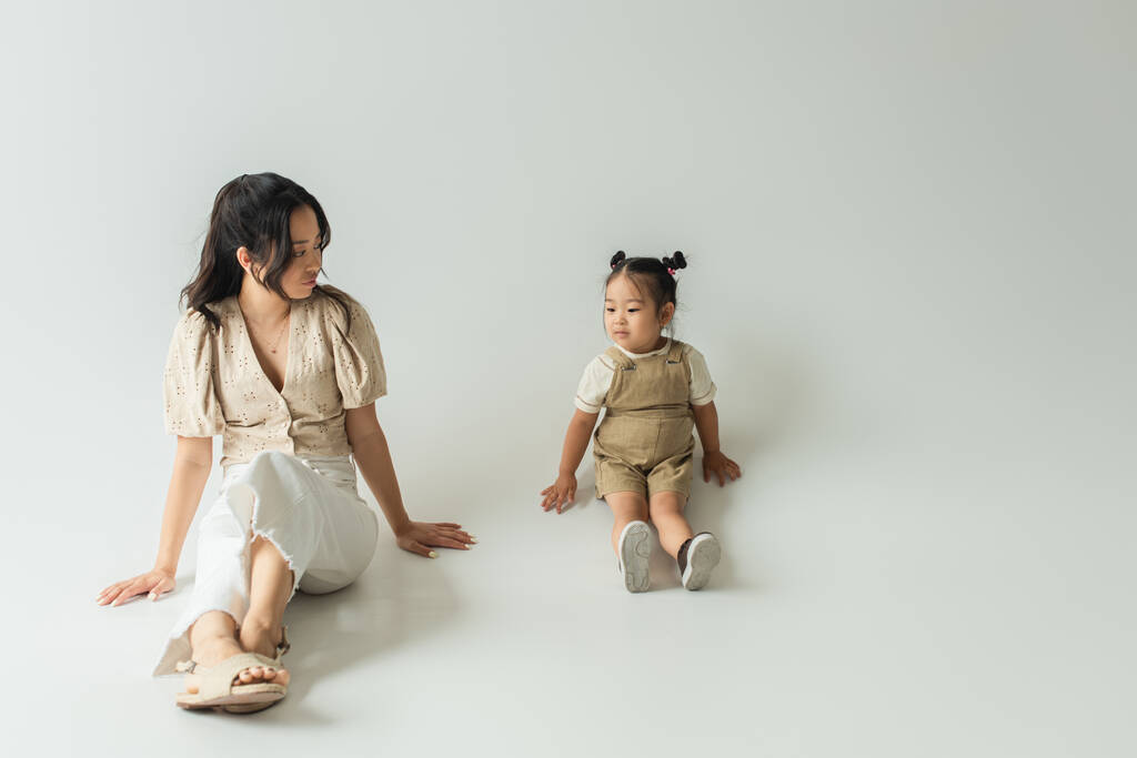 volledige lengte van aziatische moeder zitten en kijken naar peuter dochter op grijs - Foto, afbeelding