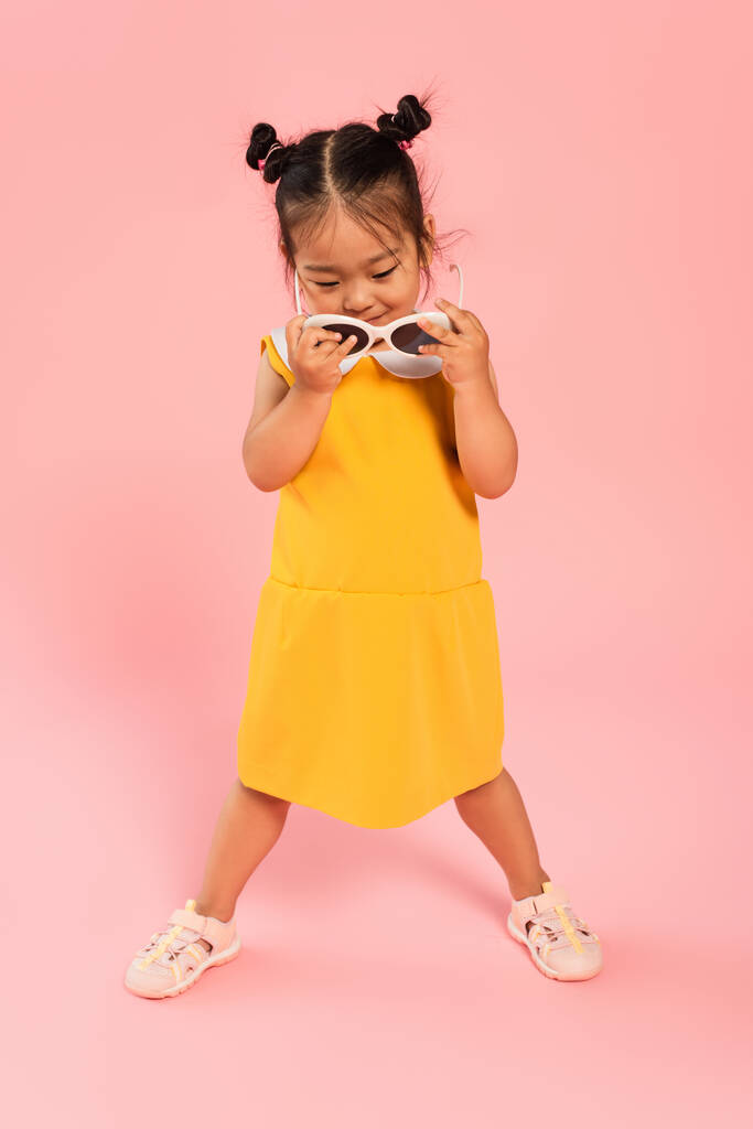 piena lunghezza di asiatico bambino in abito giallo che tiene occhiali da sole su rosa - Foto, immagini