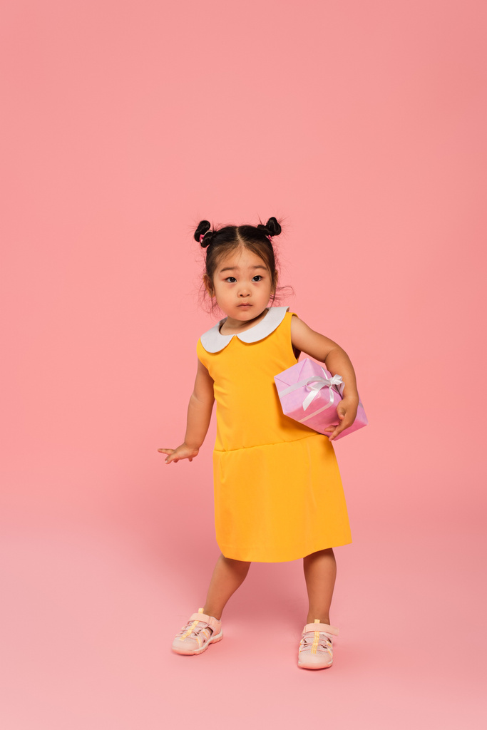 piena lunghezza di asiatico bambino in giallo abito holding presente sopra la testa su rosa - Foto, immagini