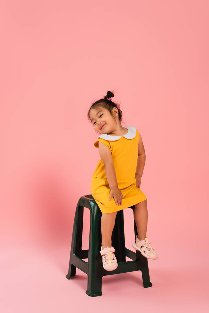 volle Länge der zufriedenen asiatischen Kleinkind in gelbem Kleid sitzt auf Stuhl auf rosa - Foto, Bild