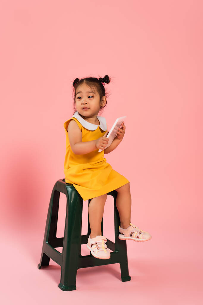 アジア系の幼児の女の子の完全な長さ黄色のドレスに座っている椅子とピンクのデジタルタブレットを使用して - 写真・画像