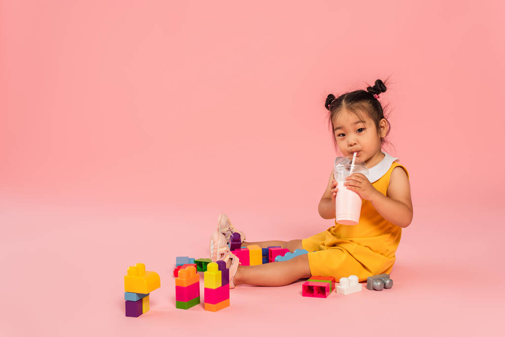 Азиатский малыш в жёлтом платье пьет вкусный молочный коктейль через солому рядом с красочными строительными блоками на розовом - Фото, изображение