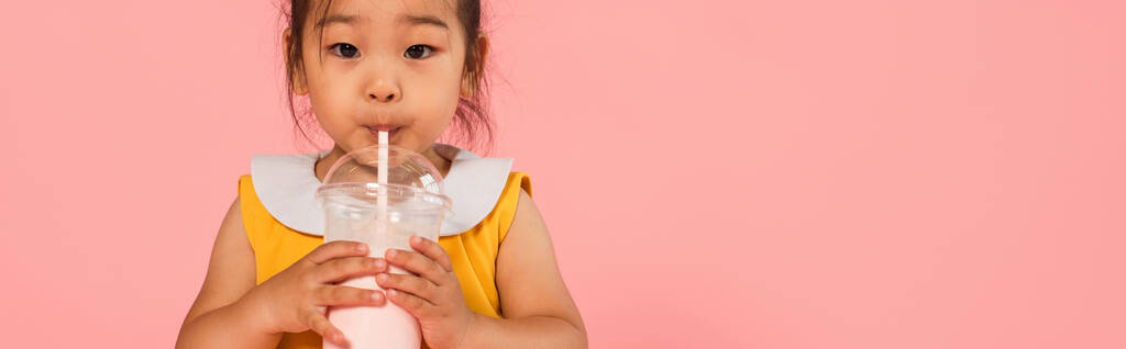Азиатский малыш в желтом платье пить вкусный молочный коктейль через солому изолированы на розовый, баннер - Фото, изображение