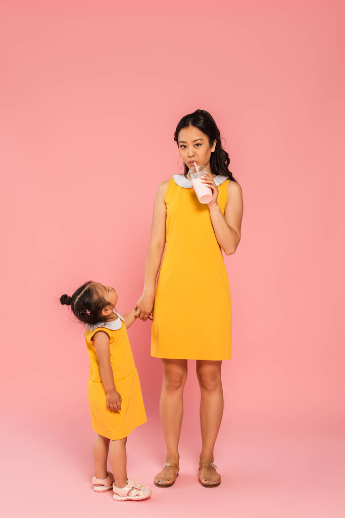 asian toddler kid in yellow dress looking at mother drinking tasty milkshake through straw on pink - Photo, Image