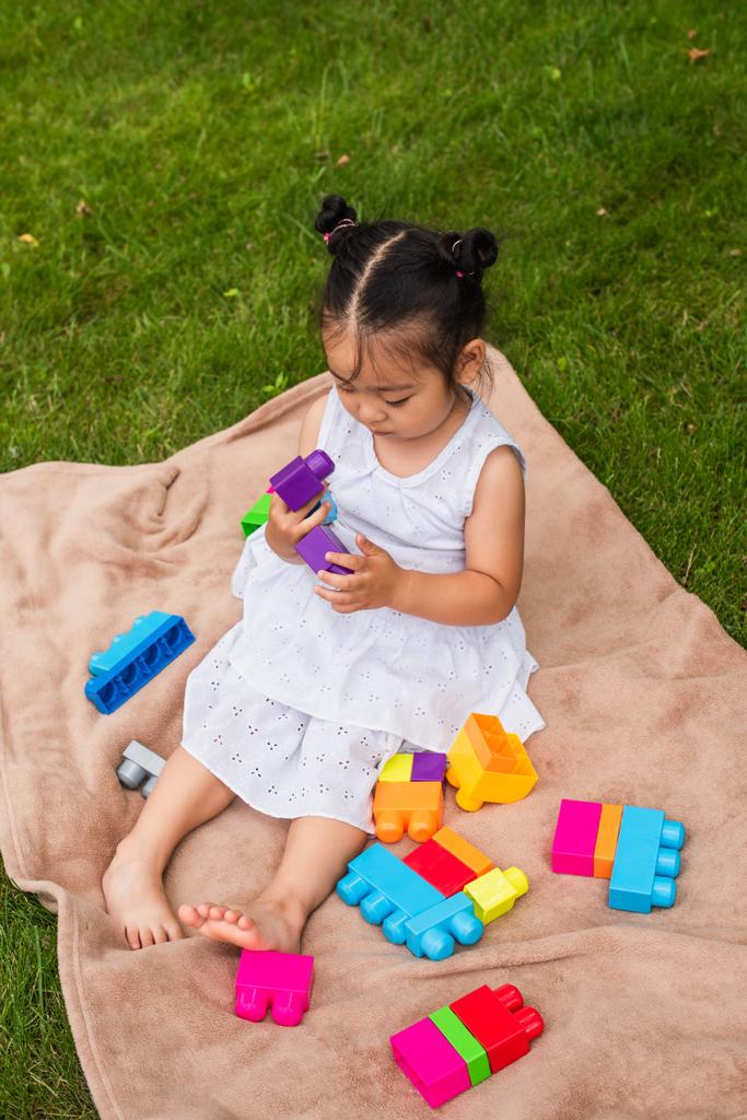 high angle view of asian child in dress playing building blocks on picnic blanket in park - Photo, Image