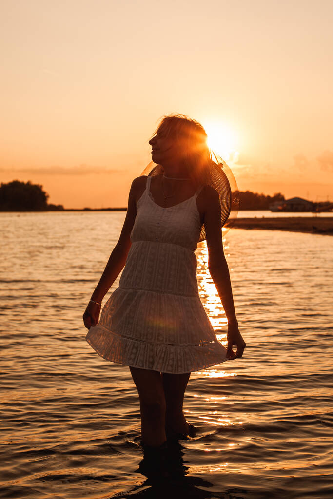 Młoda kobieta na tle zachodu słońca. Zdjęcie pięknej blondynki trzymającej obszycie białej sukienki letniej i noszącej słomiany kapelusz, pozującej w wodzie i podziwiającej przyrodę - Zdjęcie, obraz