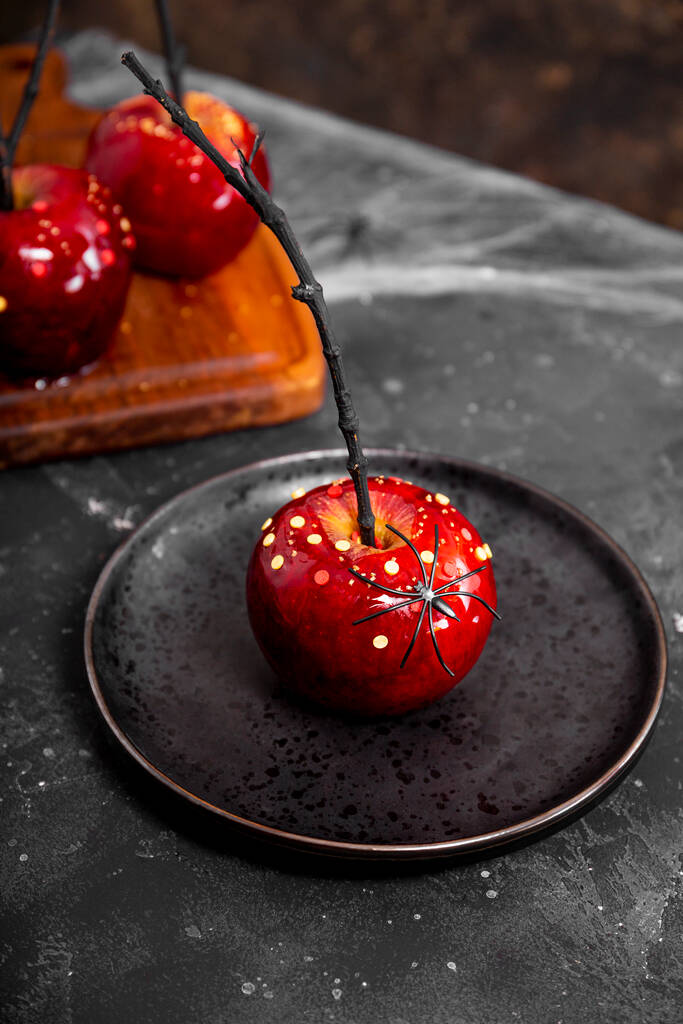 Roter Apfel in Karamell mit Zuckerdekor auf schwarzem Teller, ein origineller Leckerbissen für eine festliche Halloween-Tafel, vertikales Foto - Foto, Bild