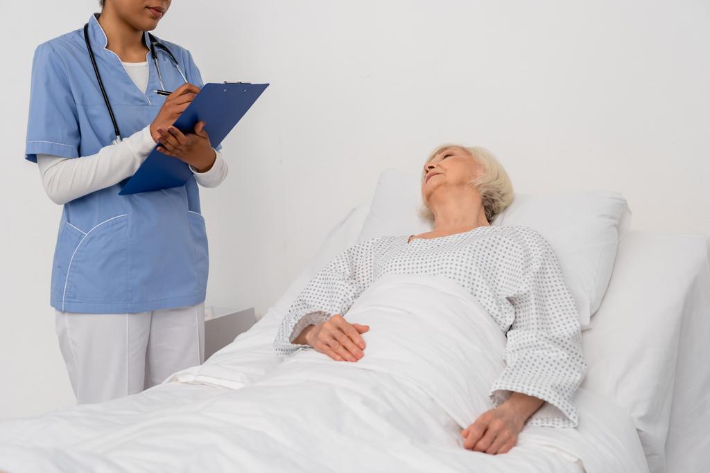 Infirmière afro-américaine écrivant sur presse-papiers près d'un patient âgé couché sur un lit d'hôpital  - Photo, image