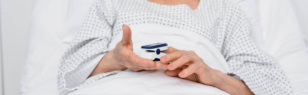 Cropped view of elderly patient holding oximeter on bed in clinic, banner  - Photo, Image