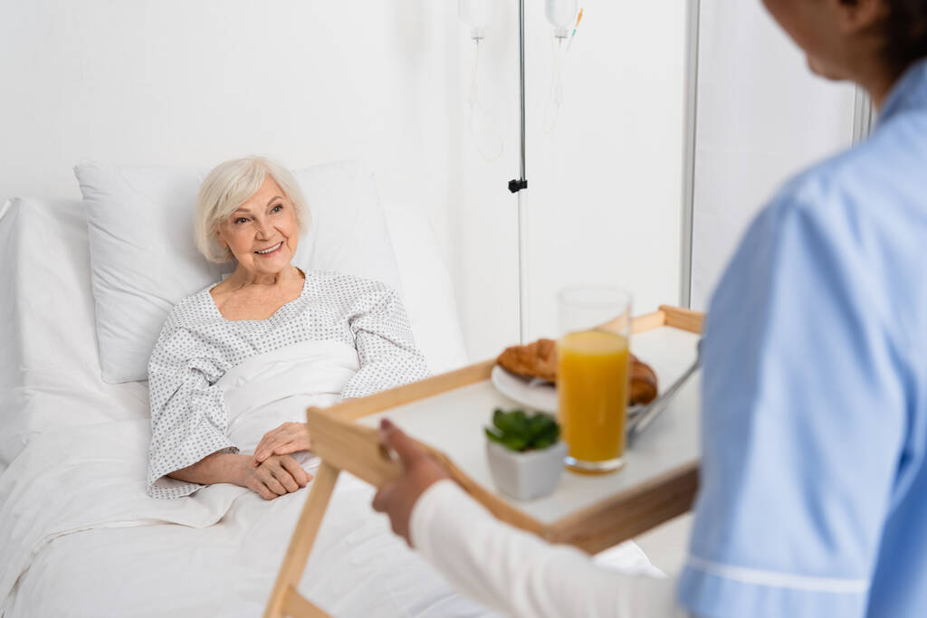 A mosolygó idősebb nő az afro-amerikai nővért nézi, miközben az ételt a homályos előtérben látja a kórházban.  - Fotó, kép