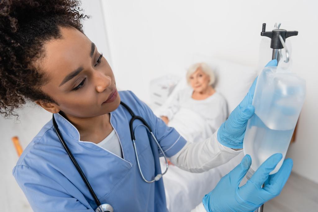 Afrikai-amerikai ápoló gumikesztyűben, aki intravénás terápiát tart a kórházi kórteremben, homályos beteg közelében.  - Fotó, kép