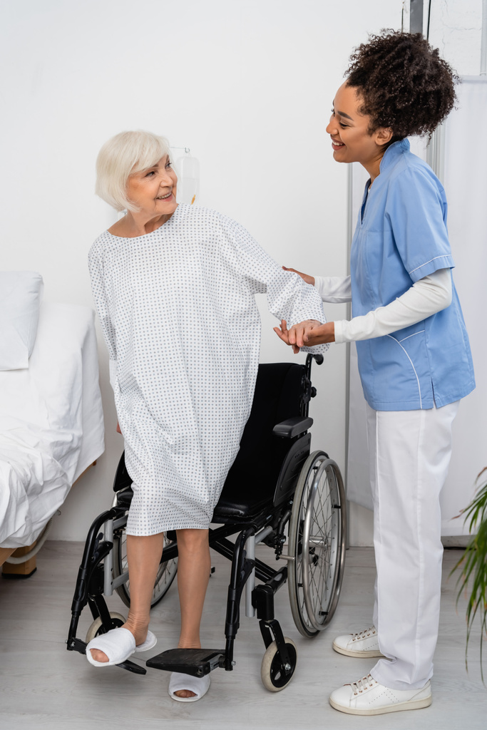 Gülümseyen Afrikalı Amerikalı hemşire klinikte tekerlekli sandalyenin yanında bir hastanın elini tutuyor.  - Fotoğraf, Görsel