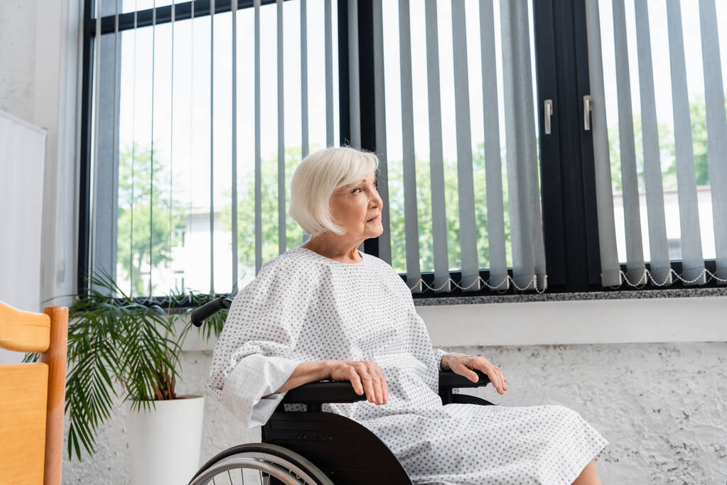 Tekerlekli sandalyedeki yaşlı kadın hastane koğuşuna bakıyor.  - Fotoğraf, Görsel
