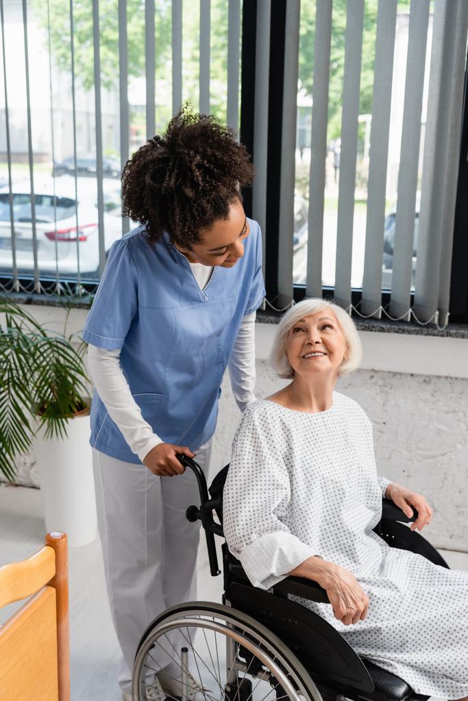 Afro-Amerikaanse verpleegster op zoek naar oudere patiënt in jurk en rolstoel  - Foto, afbeelding