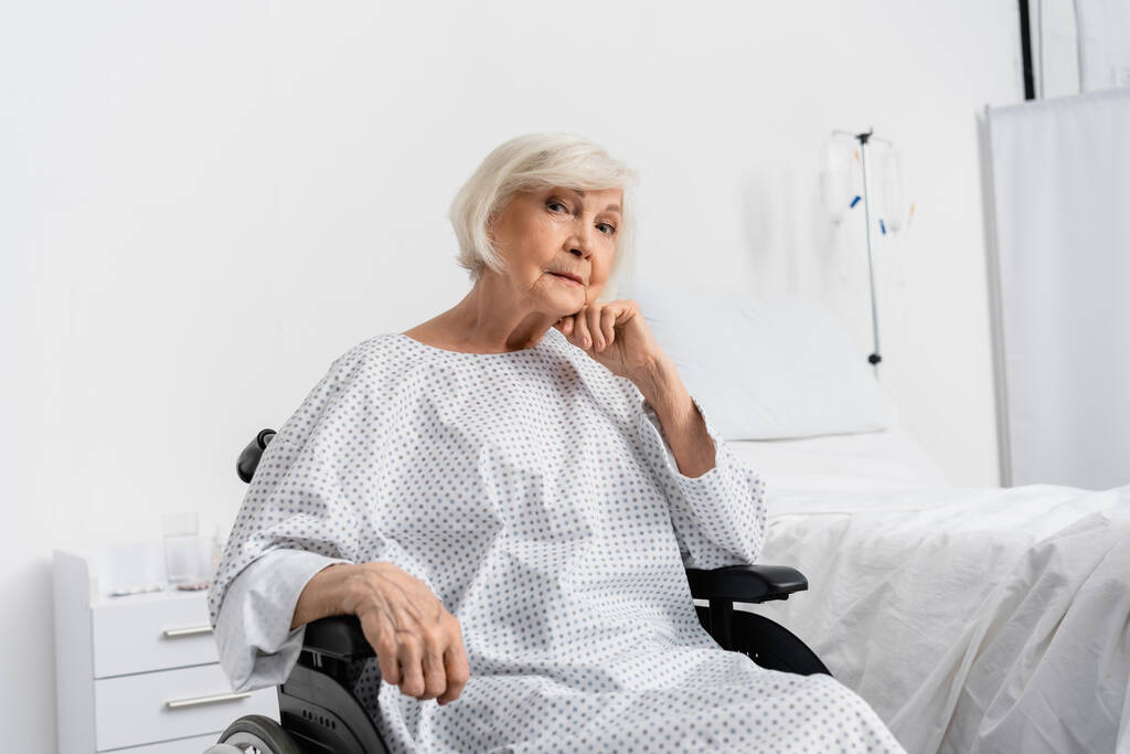 Старшая женщина в больничном халате сидит в инвалидном кресле  - Фото, изображение