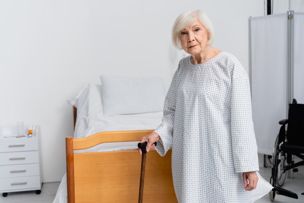 Ηλικιωμένος ασθενής με το περπάτημα ζαχαροκάλαμο κοιτάζοντας κάμερα κοντά στο κρεβάτι και αναπηρική καρέκλα  - Φωτογραφία, εικόνα