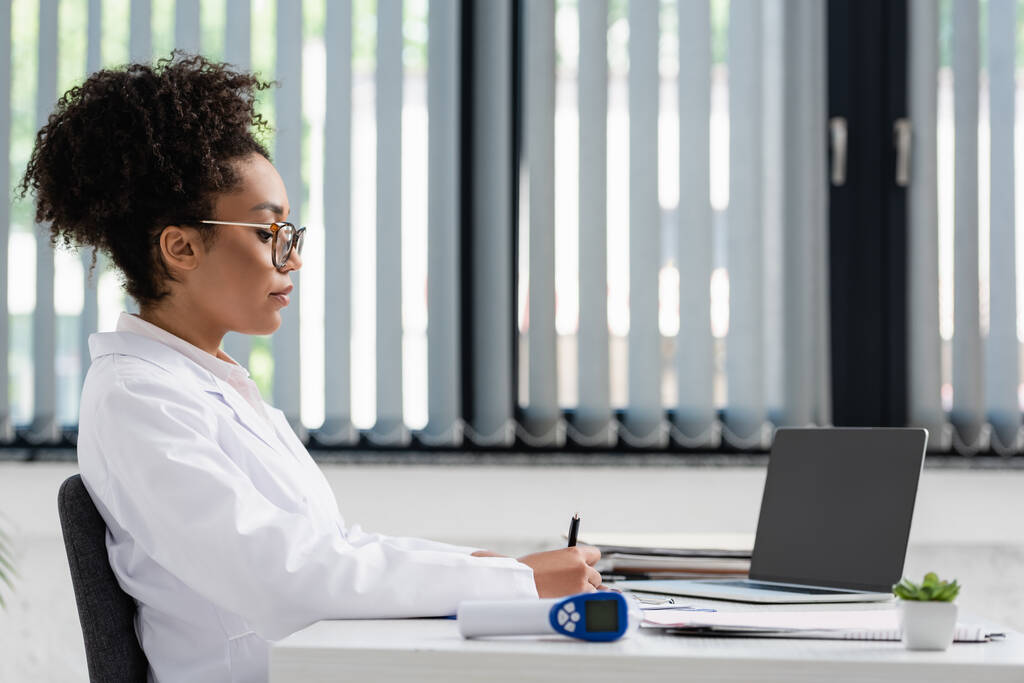 Afro-Amerikalı doktorun dizüstü bilgisayarın yanında elinde kalem tutuşu ve bulanık pirometre  - Fotoğraf, Görsel
