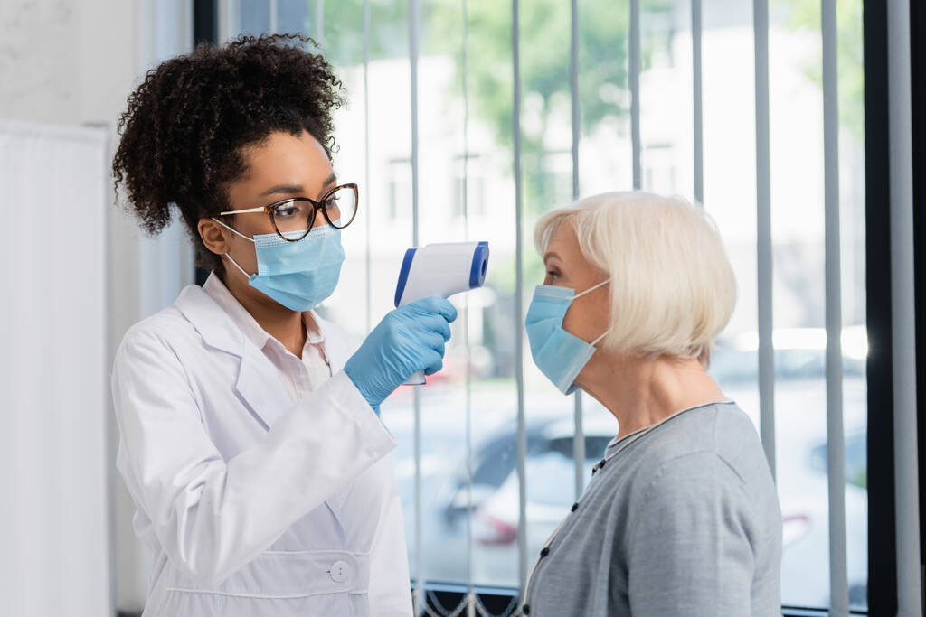 Médecin afro-américain vérifiant la température du patient dans un masque médical  - Photo, image