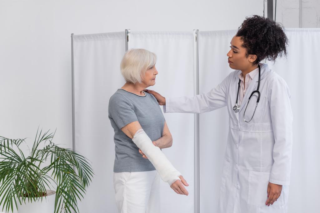 Seitenansicht eines afrikanisch-amerikanischen Arztes beruhigt Patientin mit Gipsverband am Arm in Klinik  - Foto, Bild