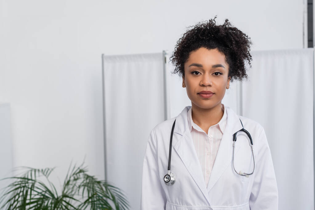 Mladý africký americký lékař v bílém plášti a stetoskop při pohledu na kameru na klinice  - Fotografie, Obrázek