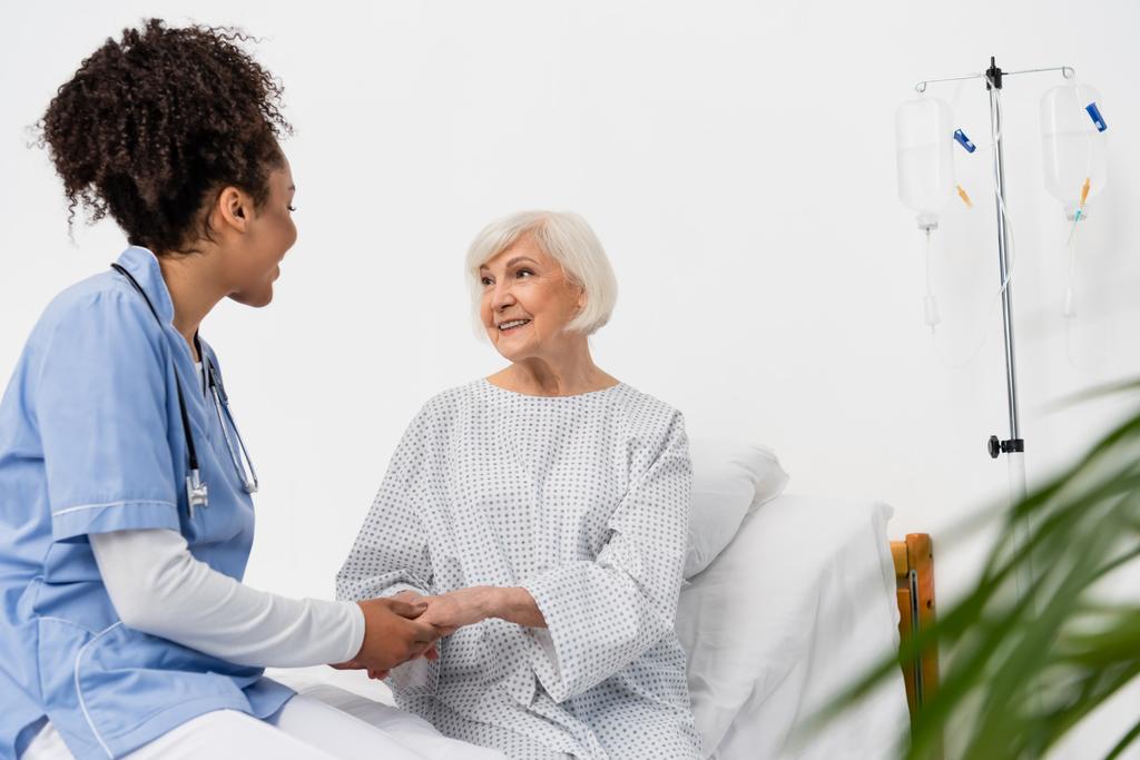 Африканська американська медсестра розмовляє зі усмішкою старшого пацієнта на лікарняному ліжку.  - Фото, зображення