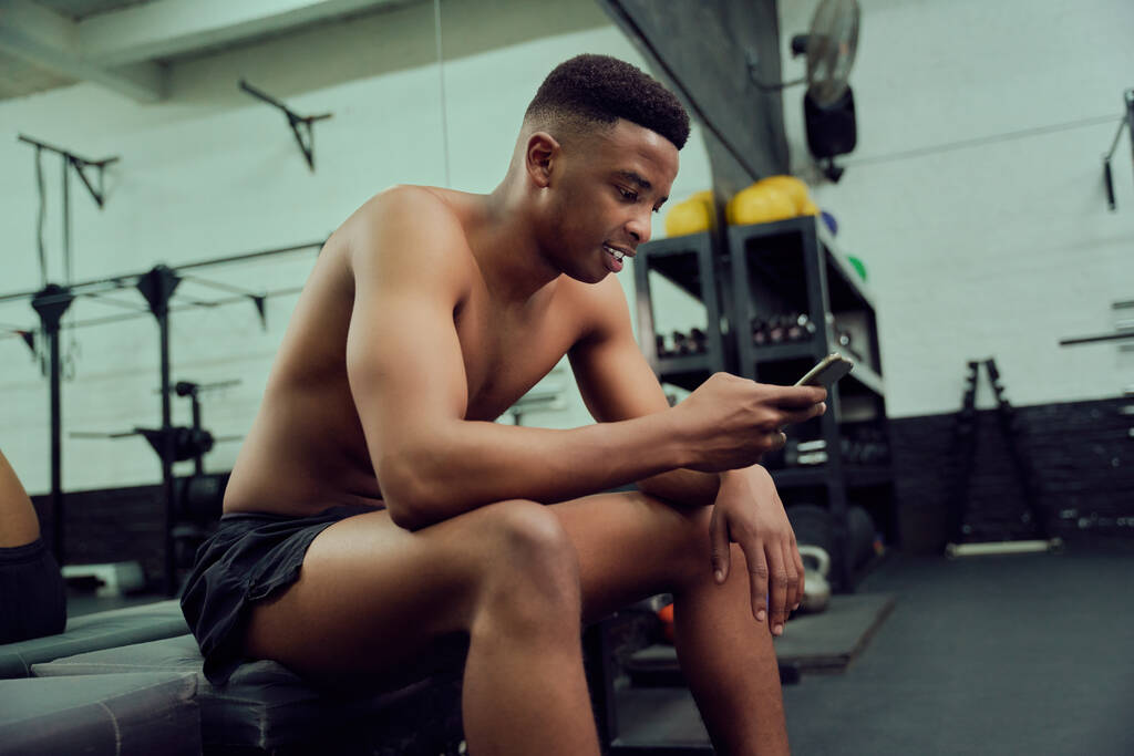 Jeune homme afro-américain regardant son téléphone pour répondre à un message en ligne. Mixte, entraîneur personnel masculin regardant son téléphone tout en s'asseyant dans la salle de gym. Photo de haute qualité  - Photo, image