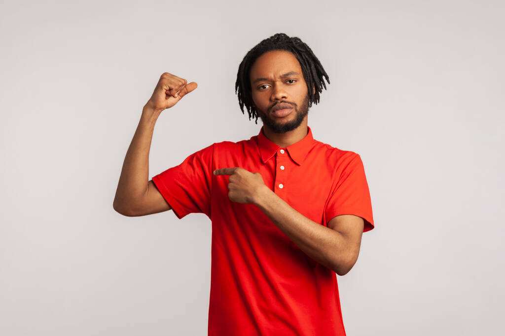Portrait of confident bearded man with dreadlocks wearing red casual style T-shirt, raising hand pointing at biceps, expressing power. Indoor studio shot isolated on gray background. - Photo, Image