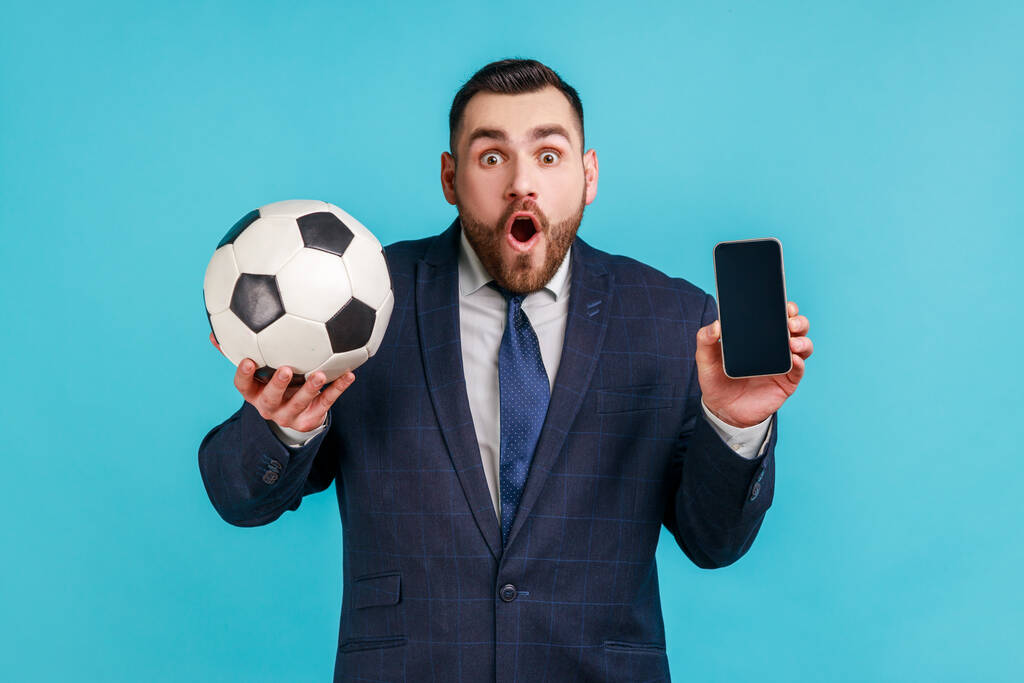 Sorprendido hombre sorprendido con barba en traje de estilo oficial sosteniendo y mostrando el teléfono inteligente pantalla negra y pelota de fútbol, apostando por el fútbol. Estudio interior plano aislado sobre fondo azul. - Foto, imagen