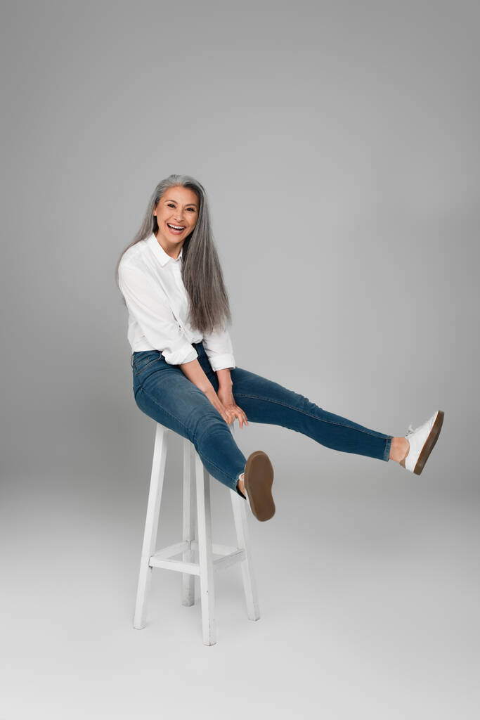 mature asian woman in blue jeans and white shirt laughing on high stool on grey background - Photo, Image