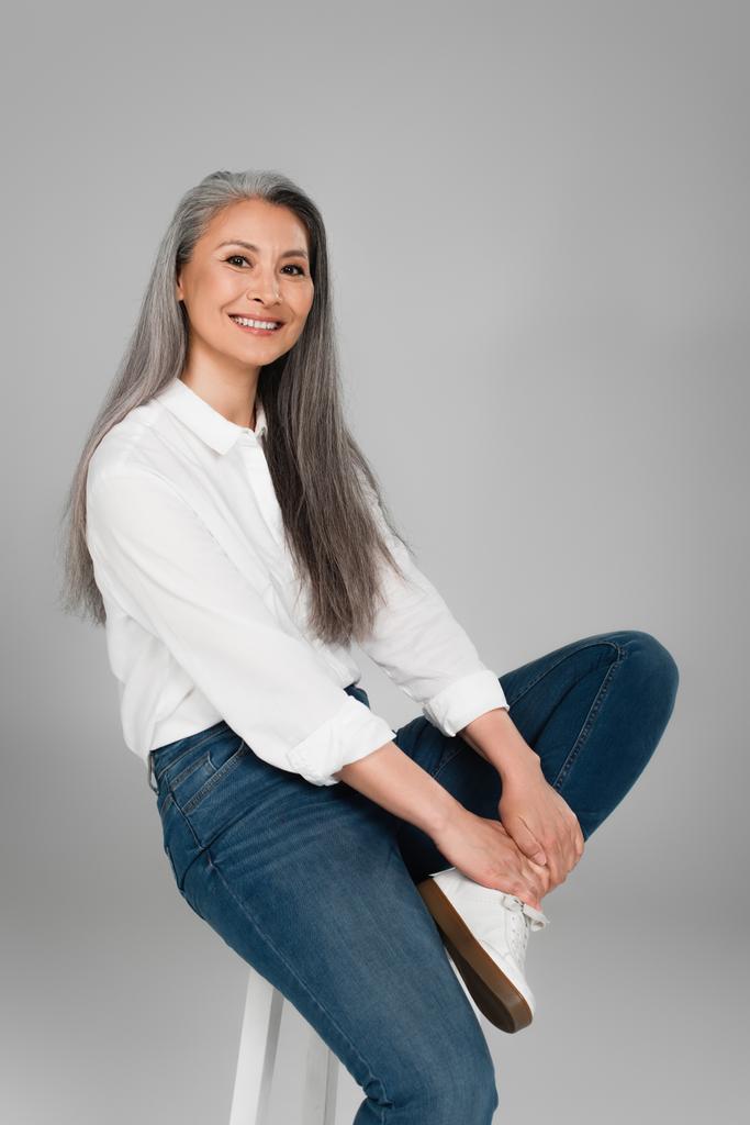 feliz asiático mujer vistiendo azul jeans y blanco camisa, sentado en taburete aislado en gris - Foto, Imagen