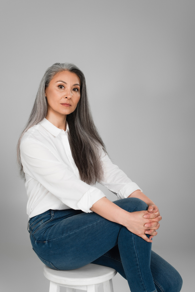 gris poil asiatique femme en blanc chemise et jeans assis sur tabouret isolé sur gris - Photo, image