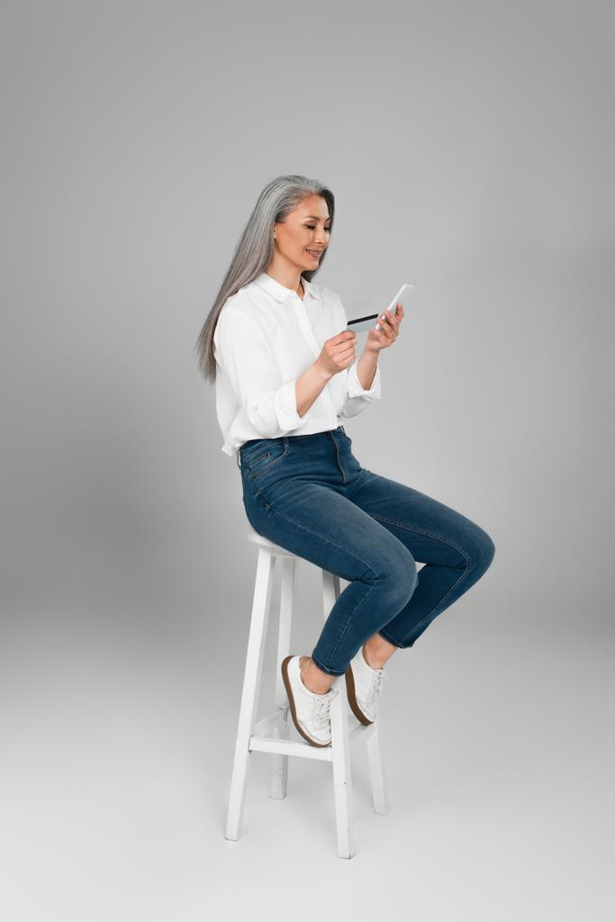 grey haired asian woman in jeans and shirt sitting on high stool with cellphone and credit card on grey background - Photo, Image