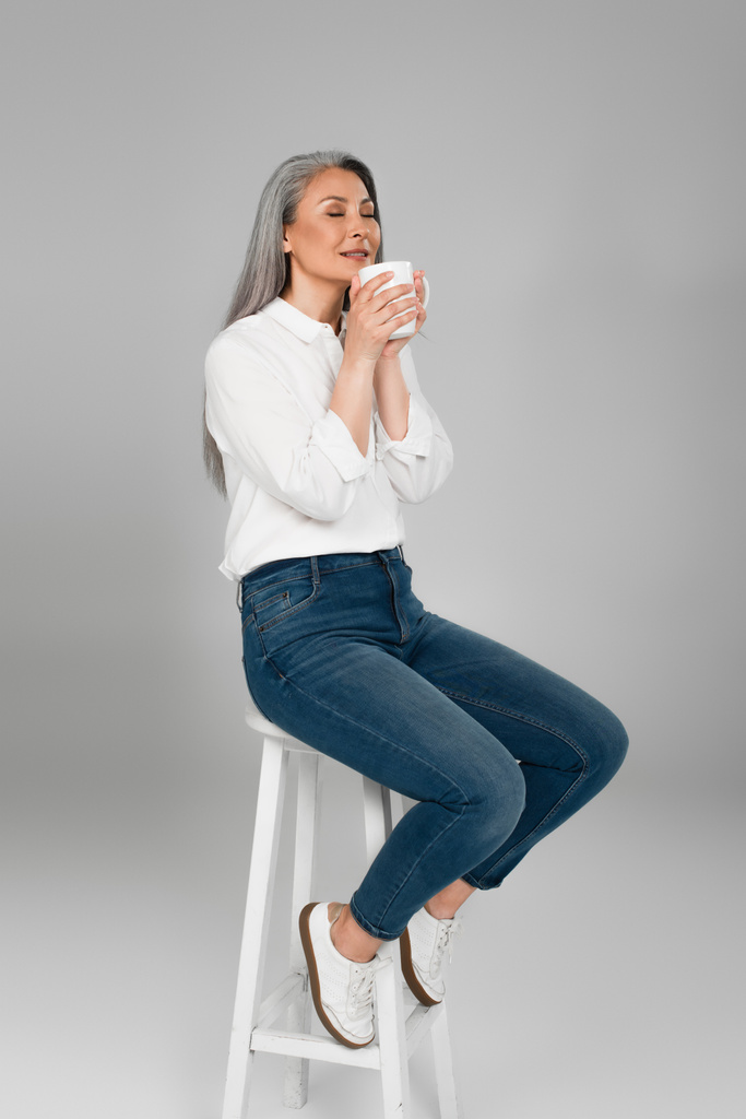 азиатская женщина средних лет в белой рубашке и джинсах сидит на стуле с чашкой ароматного чая на сером - Фото, изображение
