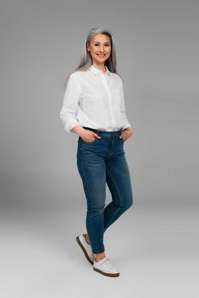 piena vista di donna asiatica con le mani in tasche di jeans blu sorridente sul grigio  - Foto, immagini
