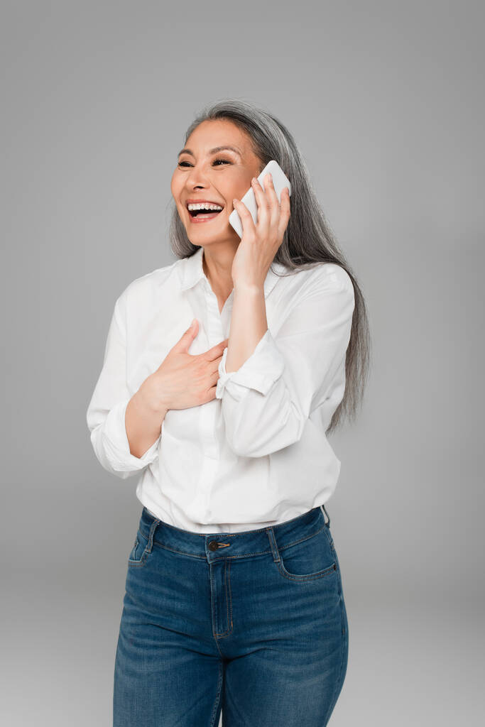 Dojrzałe Azji kobieta dotykając klatki piersiowej i śmiech podczas rozmowy na telefon komórkowy odizolowany na szary - Zdjęcie, obraz