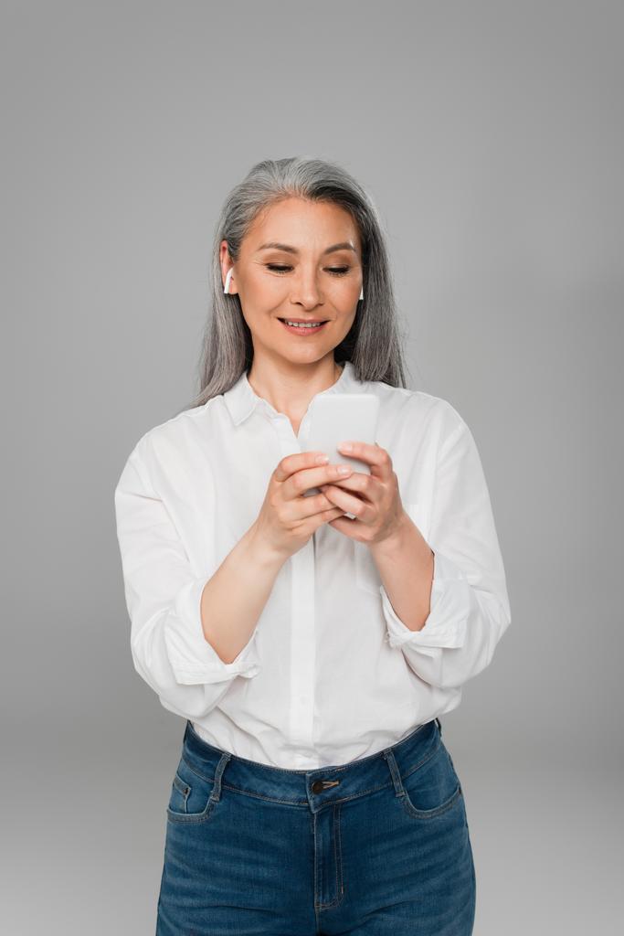 positive asiatische Frau in Kopfhörern mit Smartphone isoliert auf grau - Foto, Bild