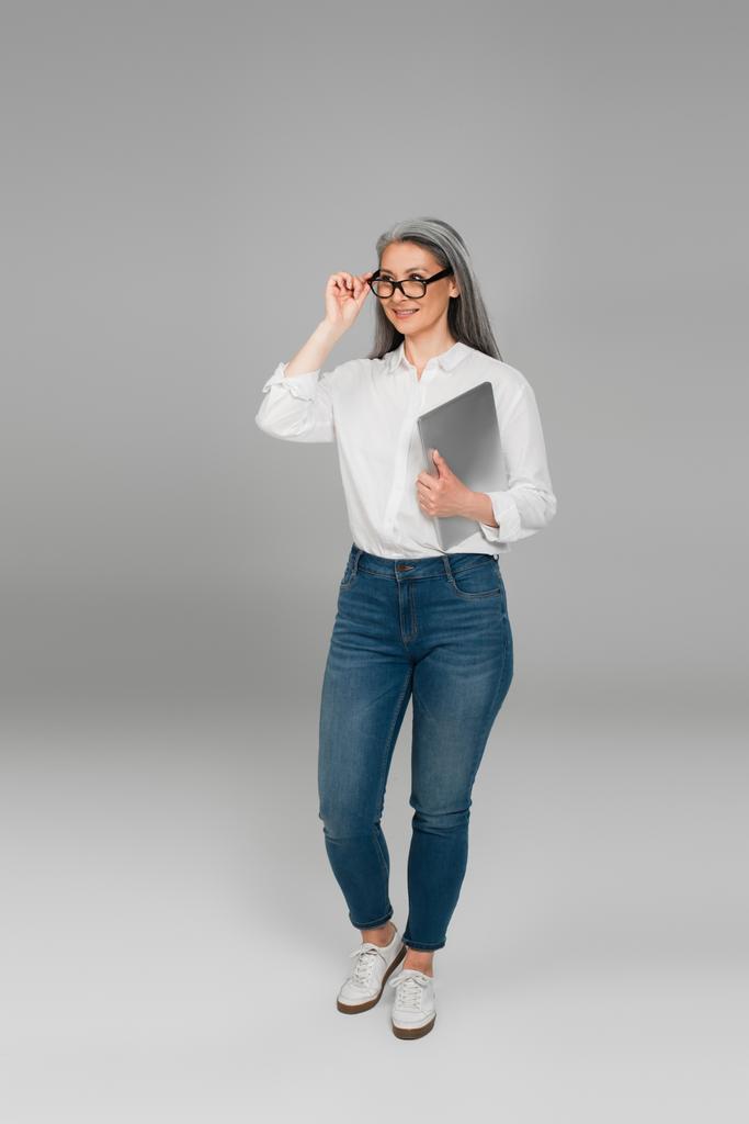 mujer asiática positiva con portátil ajustando las gafas mientras mira hacia otro lado sobre fondo gris - Foto, Imagen