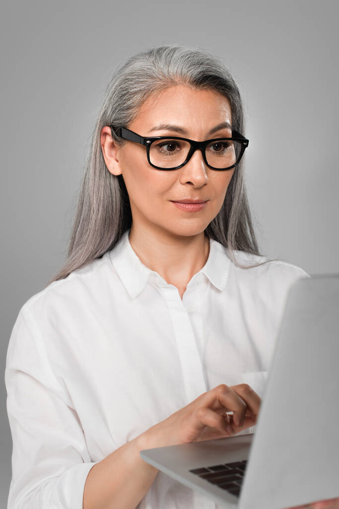 白いシャツを着た成熟したアジア系女性とグレーで隔絶されたノートパソコンにタイプするスタイリッシュな眼鏡 - 写真・画像