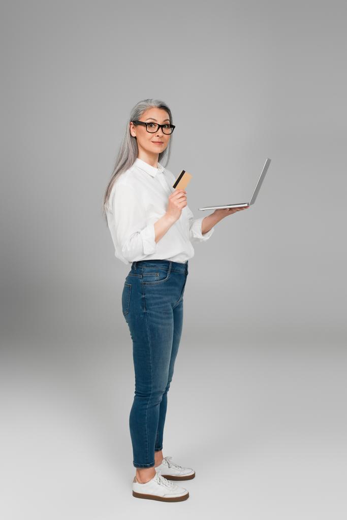 зрелая азиатка в джинсах и белой рубашке стоит с кредитной картой и ноутбуком на сером фоне - Фото, изображение