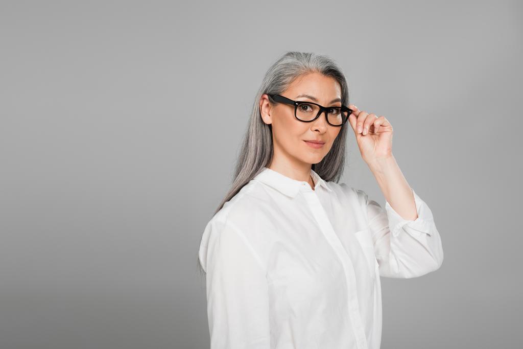 middle aged asian woman in white shirt looking at camera while touching eyeglasses isolated on grey - Photo, Image