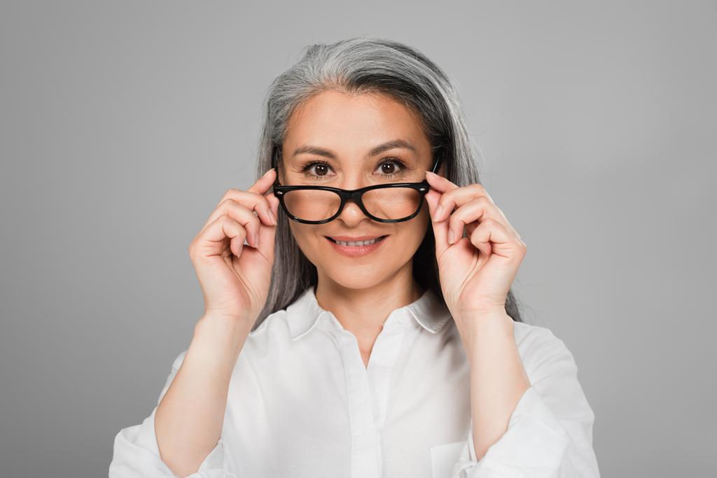 mature asiatique femme en blanc chemise décoller lunettes tout en souriant à caméra isolé sur gris - Photo, image
